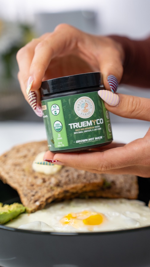 Mandi holding an Urban Roots Farms TrueMyco jar over avocado toast